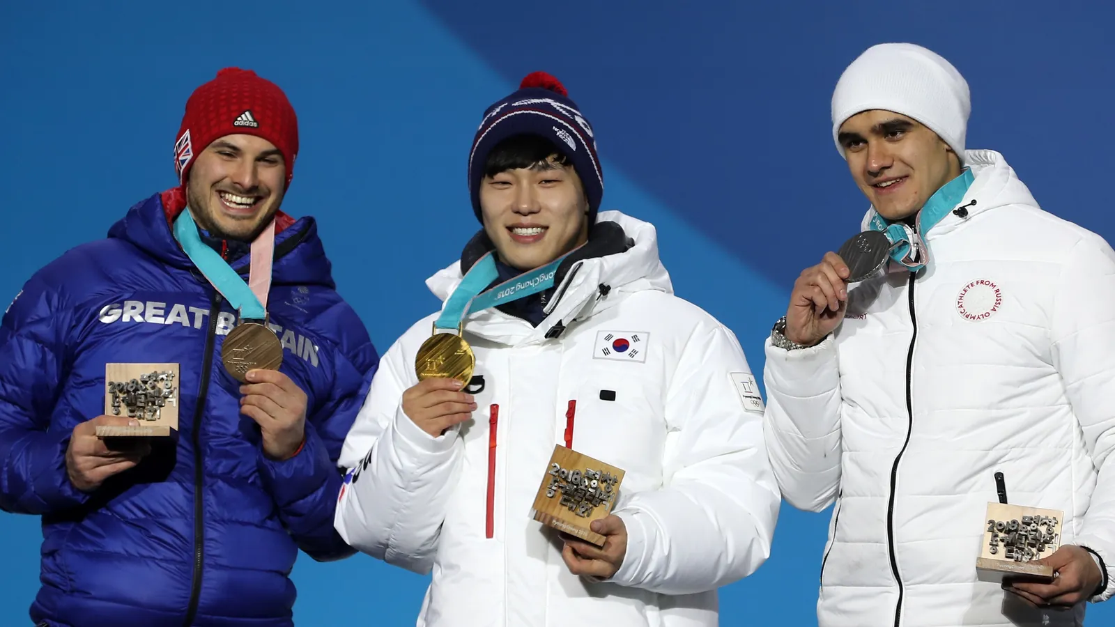 Yun Sung-bin in a white Team Korea jacket holding up his gold medal on the Olympic podium, between Dominic Parsons and Nikita Tregubov