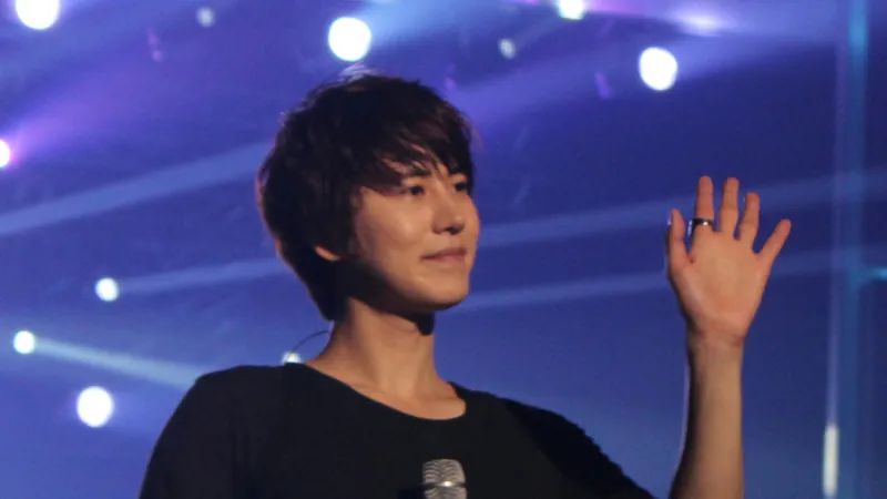 Kyuhyun in a black T-shirt waving to the crowd on a blue-lit concert stage