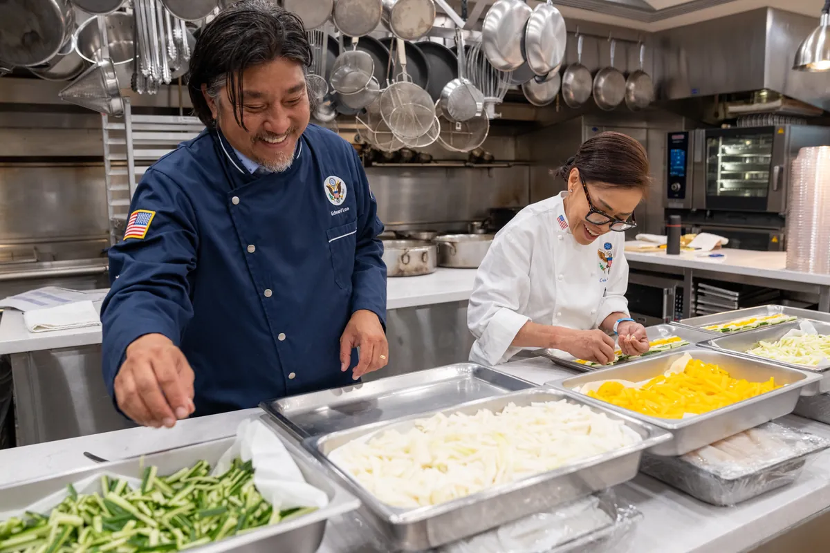 Chef Edward Lee in a navy chef jacket preparing trays of food beside Executive Chef Cristeta Comerford in the White House kitchen